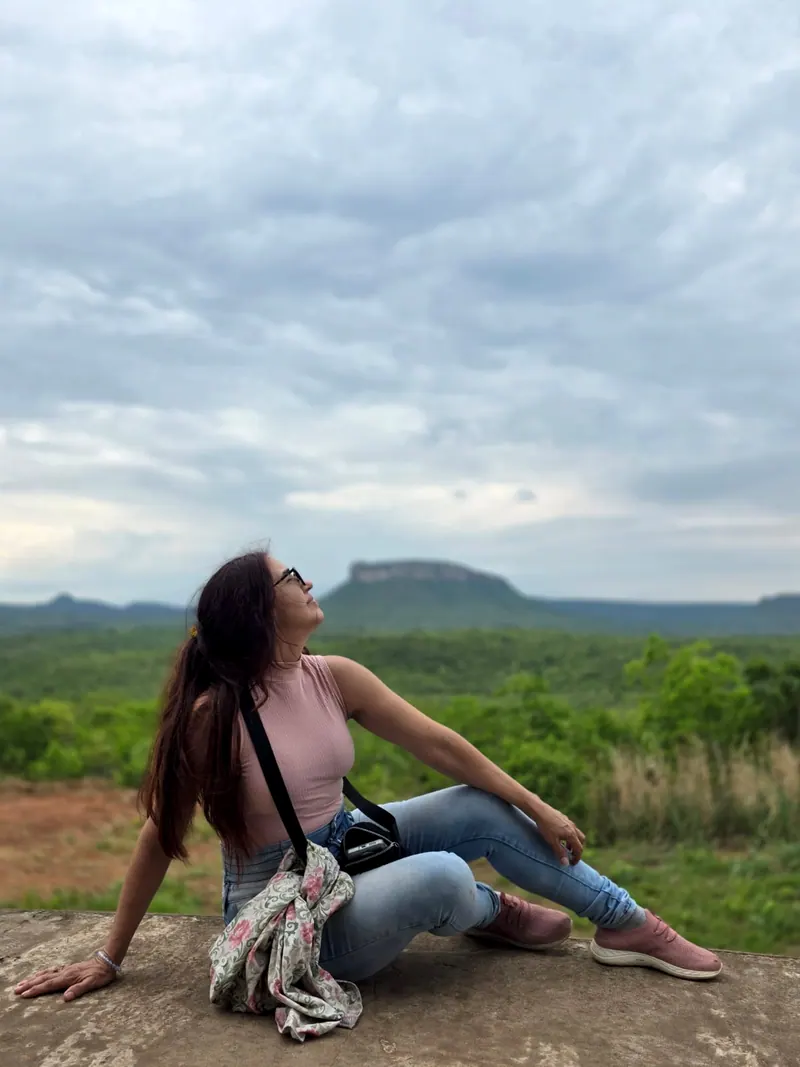 Graziela Bartiê sentada numa laje de pedra, olhando para o céu, com um morro de topo reto e o mato verde ao fundo