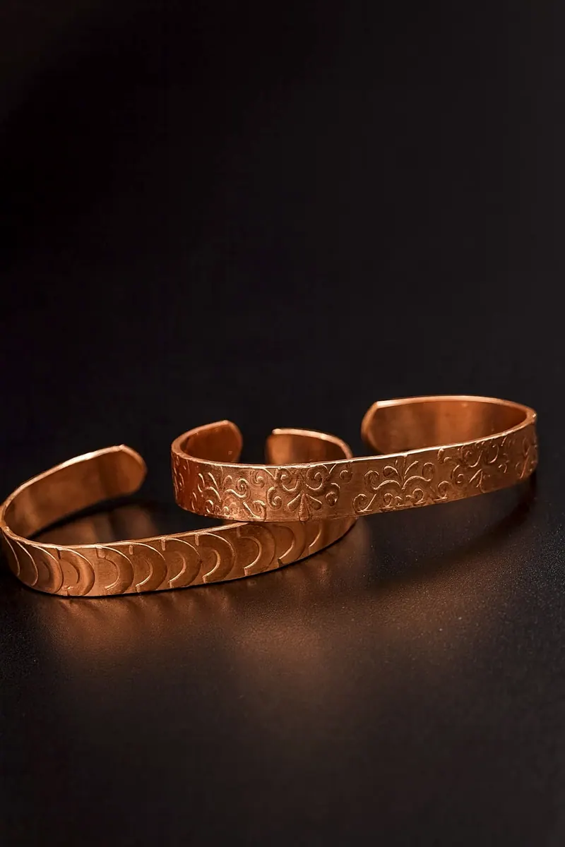 Duas pulseiras de cobre sobre fundo escuro: uma com meias-luas em relevo e outra com arabescos